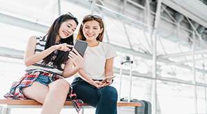 Two young Asian women sitting on a bench, engrossed in their phones, searching for airport lounges or airport experiences